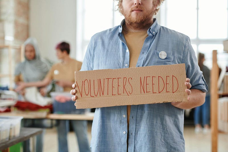 A man holding up a sign which says: Volunteers needed. Photo from Julia M Cameron on pexels.com